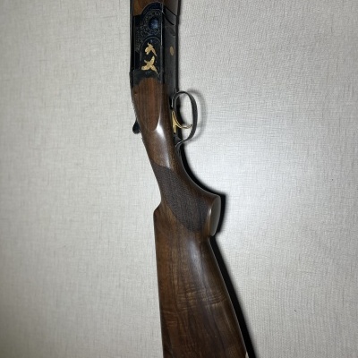 Espingarda com coronha em madeira castanha e armação preta com detalhes dourados encostada numa parede clara
