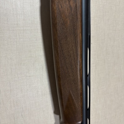 Parcial de espingarda com empunhadura em madeira decorada e cano preto junto a parede bege.