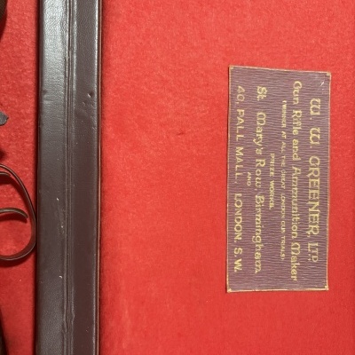 Estojo vermelho com etiqueta de fabricante e parte de arma de fogo com punho de madeira.