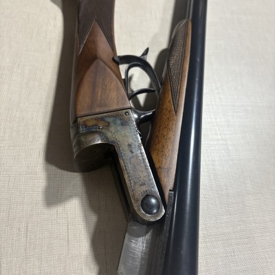 Espingarda de caça de canos duplos em metal negro e madeira com textura quadriculada, mostrada aberta.