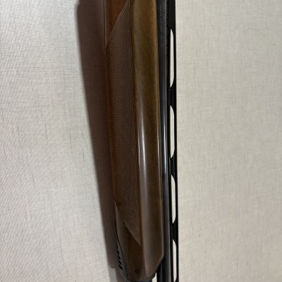 Parte da coronha e cano de uma espingarda com madeira castanha e metal preto, sobre fundo bege.