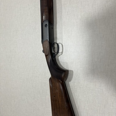 Espingarda com coronha de madeira escura e acabamento polido encostada em parede branca