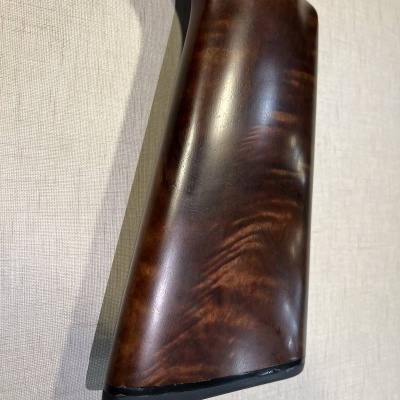 Cabo de espingarda em madeira castanha polida com padrão natural próximo a uma parede bege texturizada