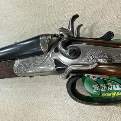 Espingarda antiga com canos em metal e coronha em madeira escura sobre superfície bege.