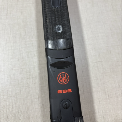 Parte de espingarda Beretta 688 em madeira e metal preto com marca vermelha sobre fundo cinzento claro.