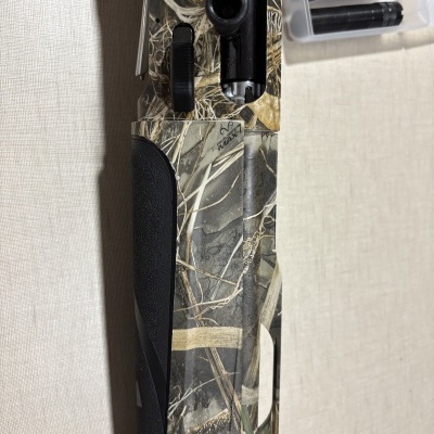 Espingarda camuflada RealTree Max-7 com três silenciadores negros numa caixa transparente ao lado.