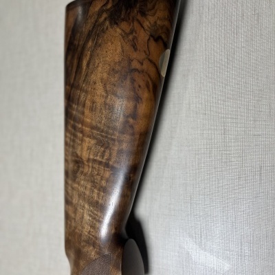 Coronha de espingarda em madeira castanho escuro com padrão natural, apoiada numa superfície cinza clara