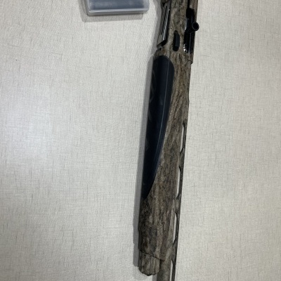 Espingarda camuflada com caixa plástica transparente e acessório sobre superfície clara