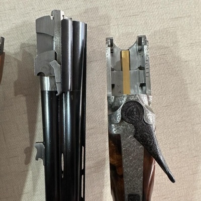 Espingarda de canos duplos desmontada com canos pretos e empunhadura em madeira sobre superfície bege