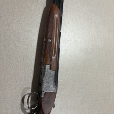 Espingarda de canos duplos com madeira castanha e metal prateado com gravações, em fundo bege.