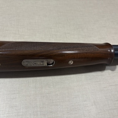 Espingarda de cano duplo com madeira castanha entalhada e detalhes metálicos sobre superfície bege.