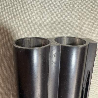 Cano duplo de arma de fogo em metal preto com acabamento mate em fundo cinza claro texturizado.
