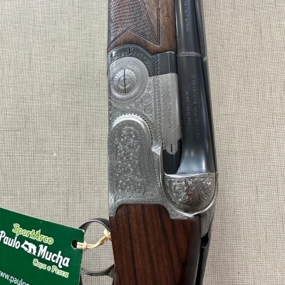 Espingarda de cano duplo com madeira castanha e detalhes gravados, com etiqueta verde pendurada.