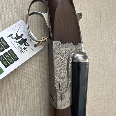 Espingarda de caça com madeira castanha e metal gravado, etiqueta VISPORT ARCO branca pendurada, sobre fundo bege.