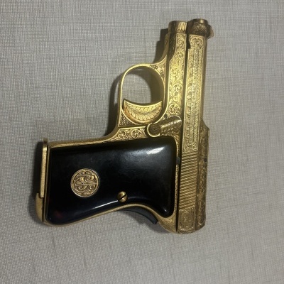 Pistola dourada com detalhes gravados e punho preto com emblema dourado sobre fundo cinza