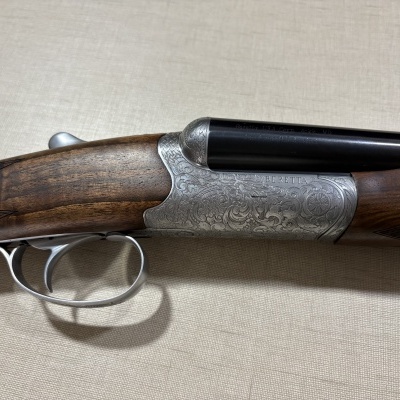 Espingarda de canos lado a lado com caixa metal gravada, coronha madeira castanha e detalhes em metal prateado.