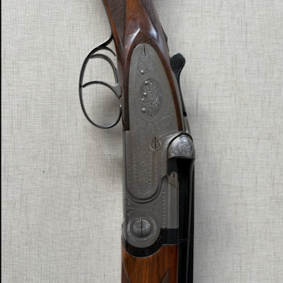 Espingarda de caça de madeira e metal com gravações ornamentais e gatilho duplo num fundo cinzento liso.