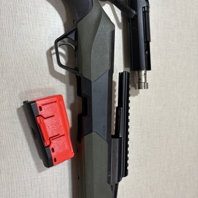 Rifle Beretta preto e verde com carregador vermelho e preto sobre fundo bege.