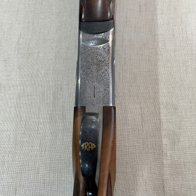 Espingarda de caça em madeira castanha e metal prateado com gravações e botão 'TRAP'.