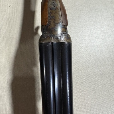 Arma de canos duplos em metal negro e madeira com gravações e inscrições sobre fundo tecido cinza claro.