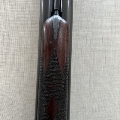 Espingarda de canos duplos com cabo de madeira escura e detalhe padrão diamante, em fundo bege.