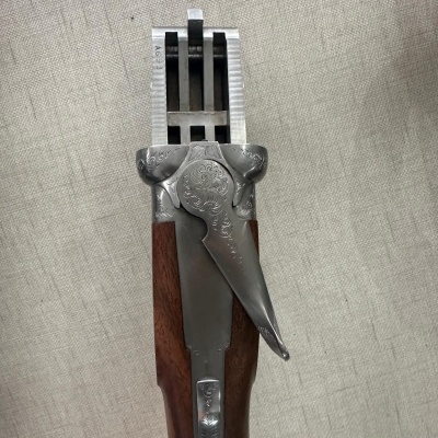 Arma de fogo de cano duplo aberta com cabo de madeira e metal gravado, sobre tecido cinza.