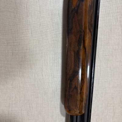 Arma de caça com cabo em madeira castanha e cano metálico preto encostada numa parede de papel de parede bege claro.