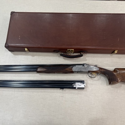 Arma de caça com coronha em madeira, canos de metal escuro e caixa rígida marrom