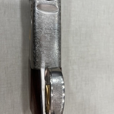 Espingarda com caixa metálica gravada e cabo de madeira sobre fundo cinza claro