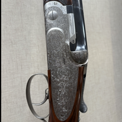 Espingarda com caixa gravada em metal prateado e madeira escura ao fundo bege