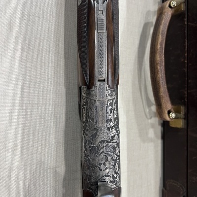 Espingarda em madeira e metal gravado sobre superfície clara, próxima de mala castanha