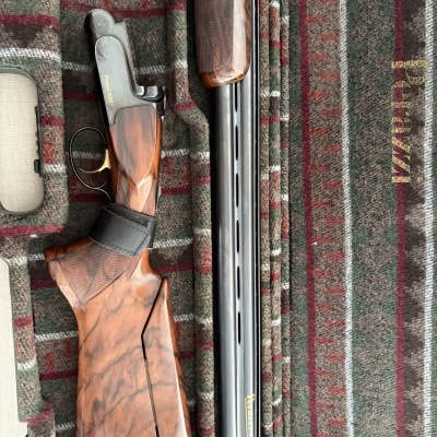 Espingarda Perazzi MX8 em madeira castanha e metal preto sobre tecido xadrez com texto