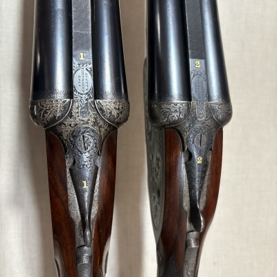 Par de espingardas com canos paralelos em metal preto, detalhes gravados e cabos de madeira castanha