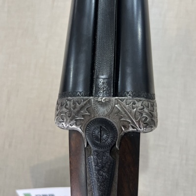 Espingarda dupla com canos negros, gravações metálicas e empunhadura em madeira escura sobre superfície clara