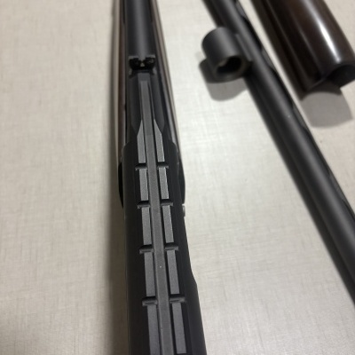 Partes metálicas pretas e madeira castanha de uma espingarda sobre superfície clara.