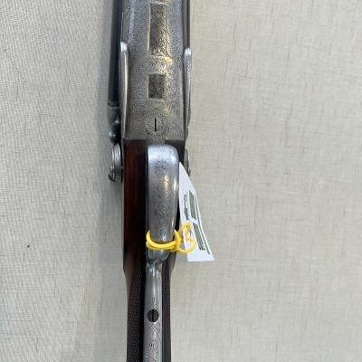 Espingarda de caça com cabo em madeira escura e metal envelhecido com etiqueta pendurada.