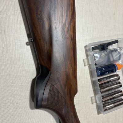 Coronha de madeira escura com acabamento brilhante e texturas, ao lado caixa transparente com ferramentas metálicas e frasco azul com tampa laranja