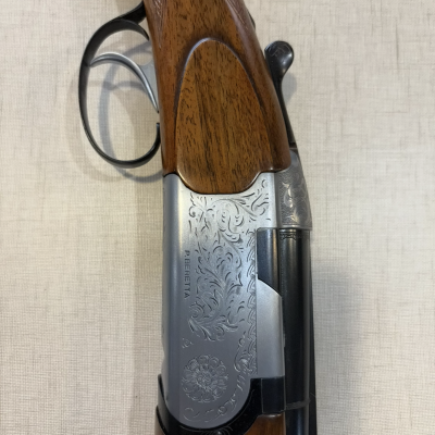Espingarda de caça com coronha em madeira e metal cravejado sobre fundo bege.