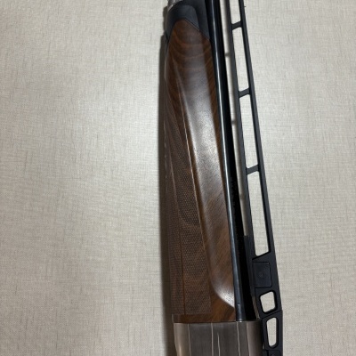 Espingarda com cano metálico escuro e empunhadura em madeira castanha com textura, sobre fundo claro.