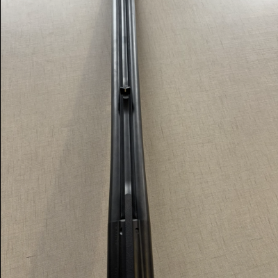 Cano duplo metálico preto de espingarda com mira vermelha sobre superfície cinzenta.