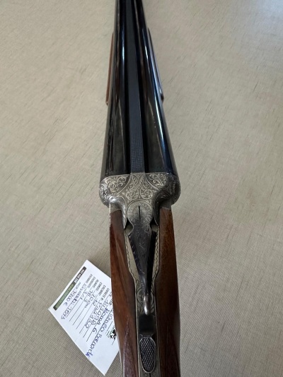 Espingarda de canos paralelos em metal preto com coronha em madeira castanha sobre superfície bege clara