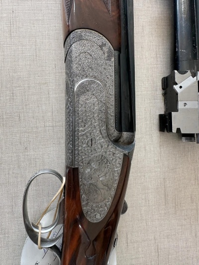 Espingarda de caça com madeira e metal gravado exibida sobre fundo claro com etiqueta