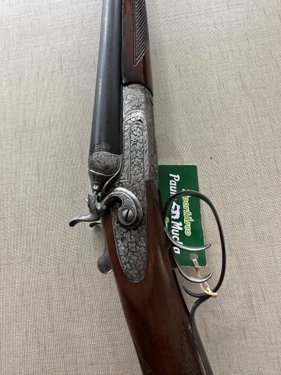 Espingarda de canos paralelos com madeira e metal entalhado, etiqueta verde com texto.