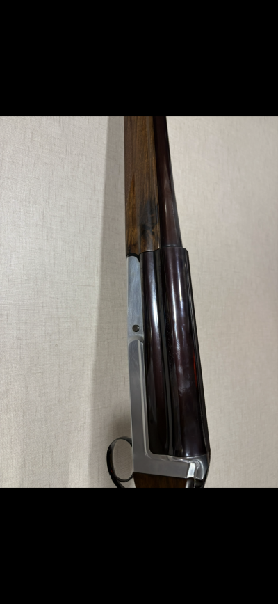 Espingarda de canos paralelos com madeira castanha e metal escuro apoiada numa parede bege.