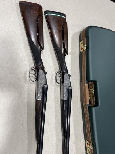 Duas espingardas de cano duplo em madeira e metal ao lado de um estojo rígido verde.