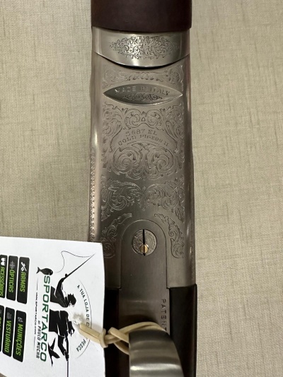 Parte superior metálica de arma com gravações e etiqueta SPORTARCO pendurada em fundo bege