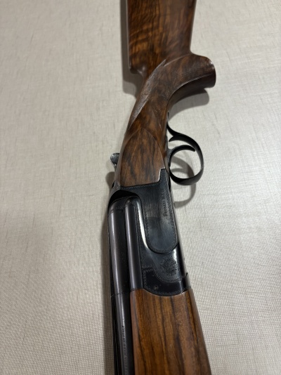 Arma de fogo Perazzi MX12 em madeira escura e metal preto sobre superfície clara.