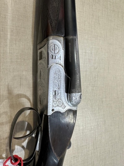 Espingarda sobreposta com metal gravado, madeira escura, gatilho duplo e fita vermelha sobre fundo bege