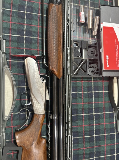 Estojo com espingarda Benelli, acessórios metálicos e manual vermelho sobre tecido xadrez.