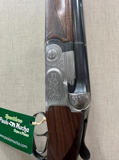 Espingarda de cano duplo com madeira castanha e detalhes gravados, com etiqueta verde pendurada.